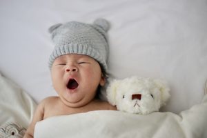 Baby yawning in bed next to a stuffed dog.
