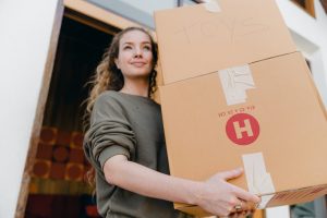 A young woman with rolled-up sleeves carrying two boxes out of her old home. 