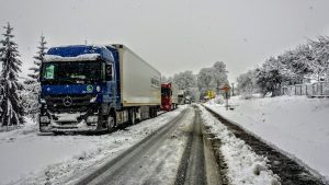 A line of cargo trucks moving slowly on a snowy road and spruce trees on the side of it.