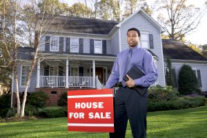 A man in a blue shirt standing in front of a two-story house next to a red 