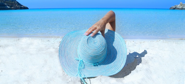 woman with a blue sun hat with a wide rim sitting on a white sandy beach with crystal blue waters