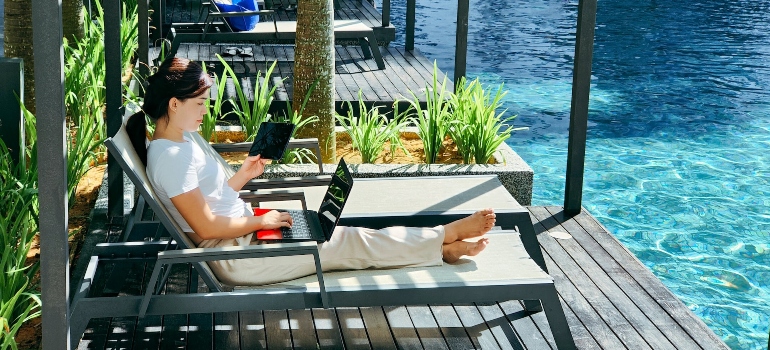 A woman sits on a deck chair by the pool and looks at a laptop and a tablet - Moving from Hallandale Beach to Hollywood FL. 