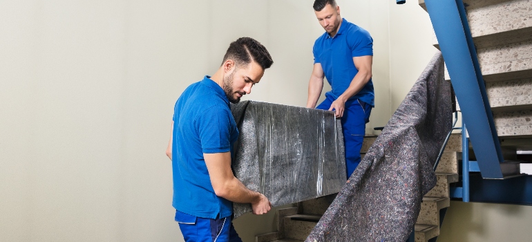 Two Young Male Movers In Uniform Carrying Furniture On Staircase - Residential movers Cedar Grove FL.