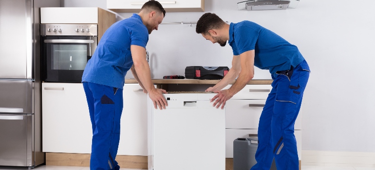 Two Young Male Movers In Uniform Placing Dishwasher In Kitchen.