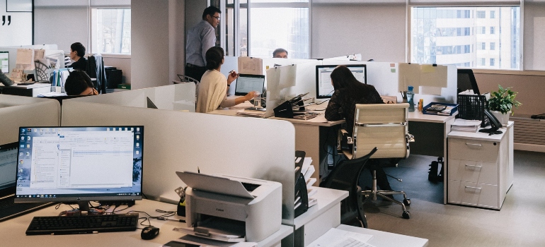 workers in a large office, office desk, computer and printer on that desk in the foreground