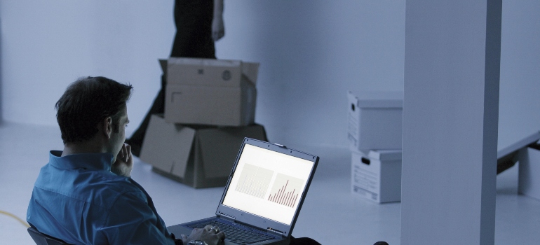 a man sits in an empty space and searches on a laptop, packed cardboard boxes can be seen behind him