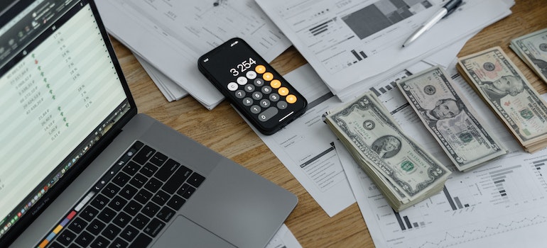 A table with laptop, dollar bills, paper and iphone on it