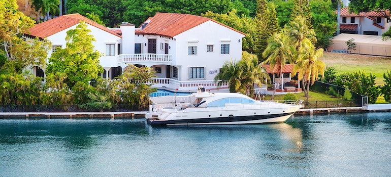 WHite waterfront house with a brown roof and lush green tress, with a boat parked in front of it
