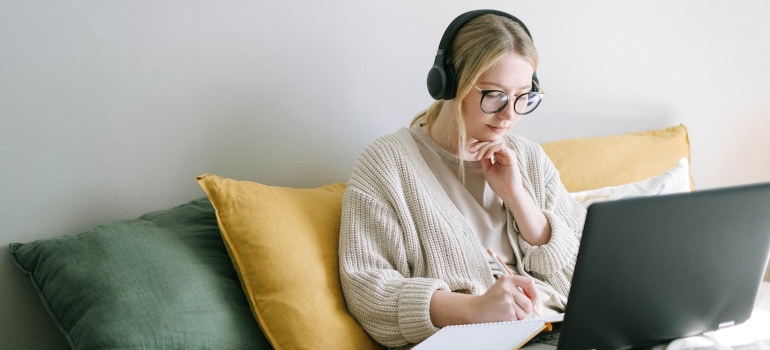 A blonde girl with glasses and headphones sitting on her bed with mustard and olive green pillows writing something in her notebook while looking at a laptop screen